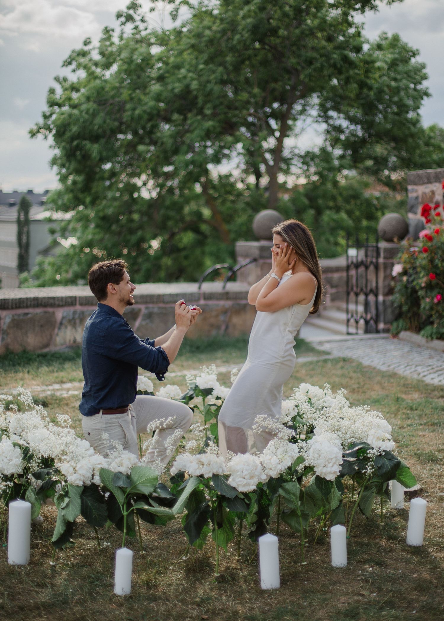 Man kneeling proposing to surprised woman surrounded by white flowers and candles in romantic garden setting.
