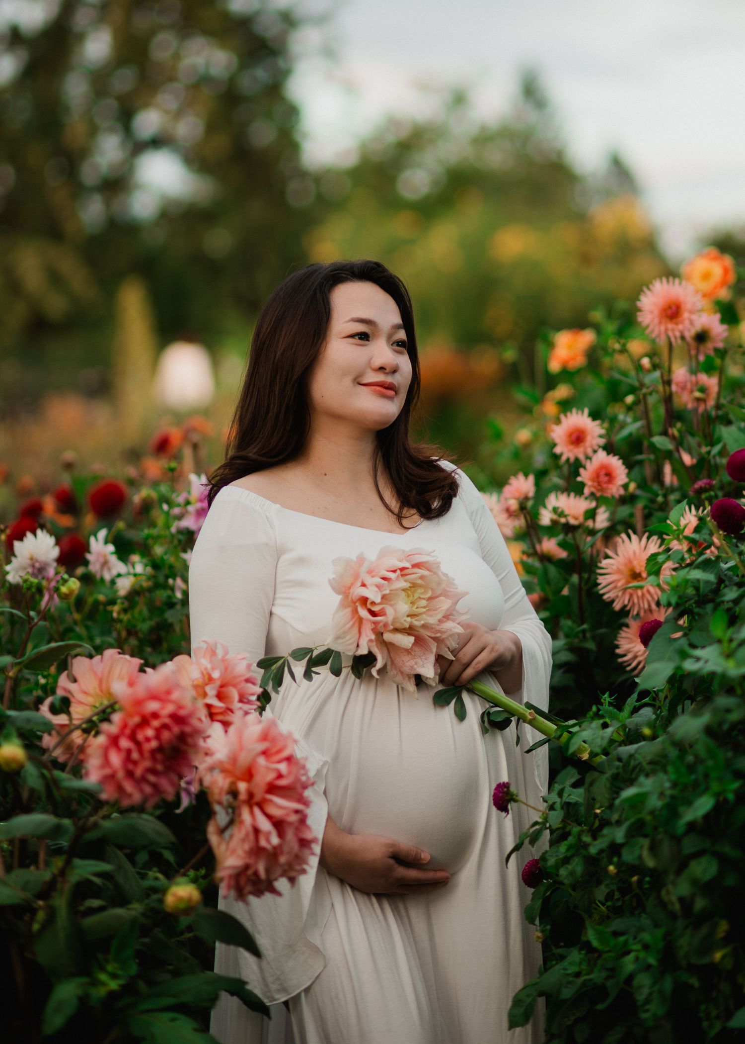 Pregnant woman in white dress holding pink dahlias in a lush flower garden.