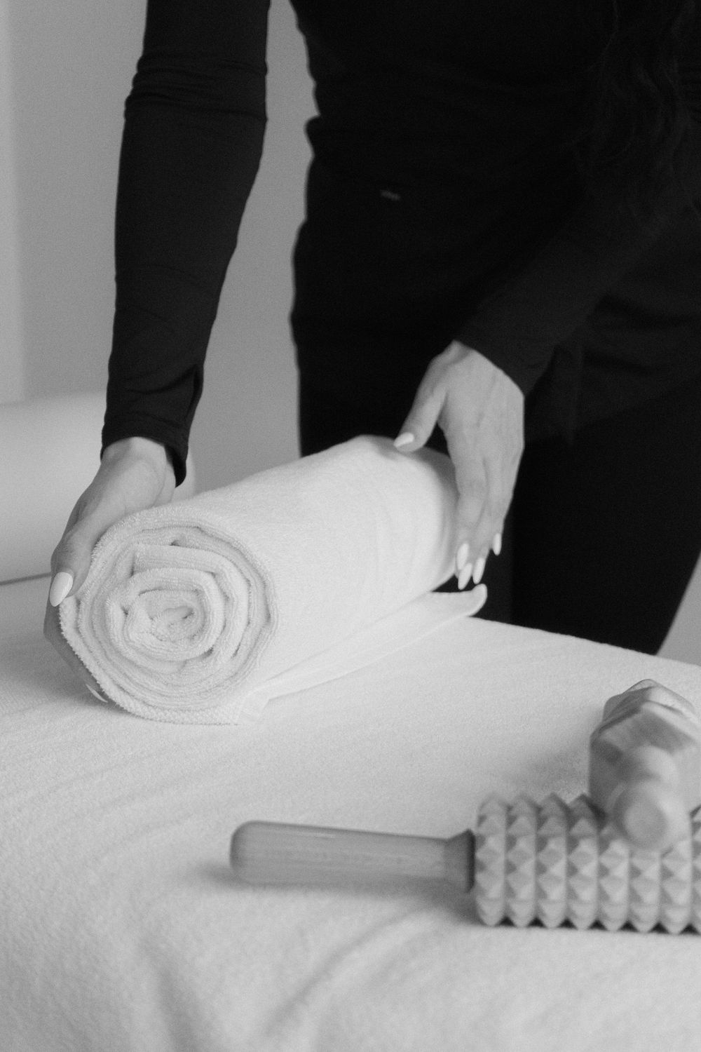Spa therapist placing a rolled white towel on a massage table with wooden massage tools nearby.