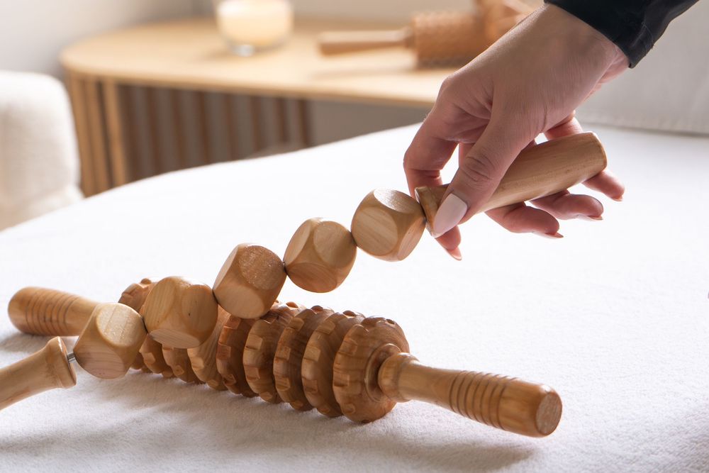 Wooden massage roller tools on white surface with hand holding a maderotherapy body sculpting instrument.