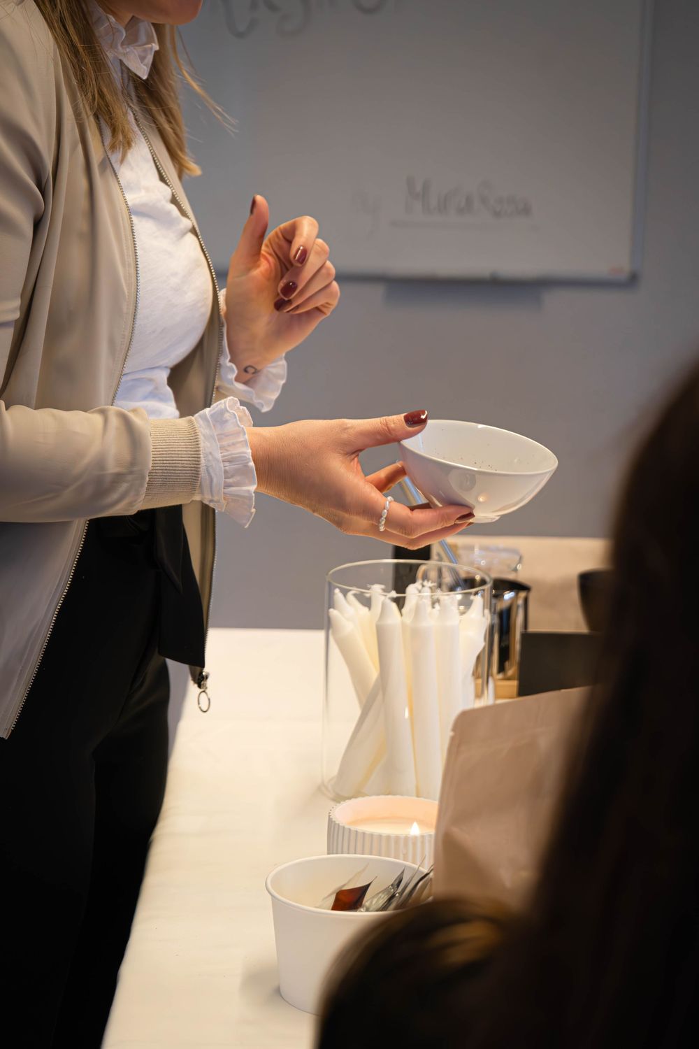 Woman holding a white bowl at a candle-making workshop with white candles and supplies on a table.