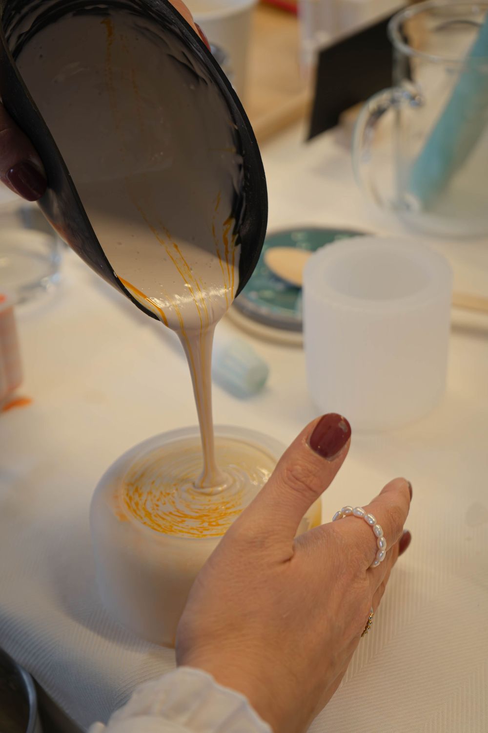 Person pouring creamy liquid into a round mold with gold swirls, crafting homemade candles or soap on a white table.