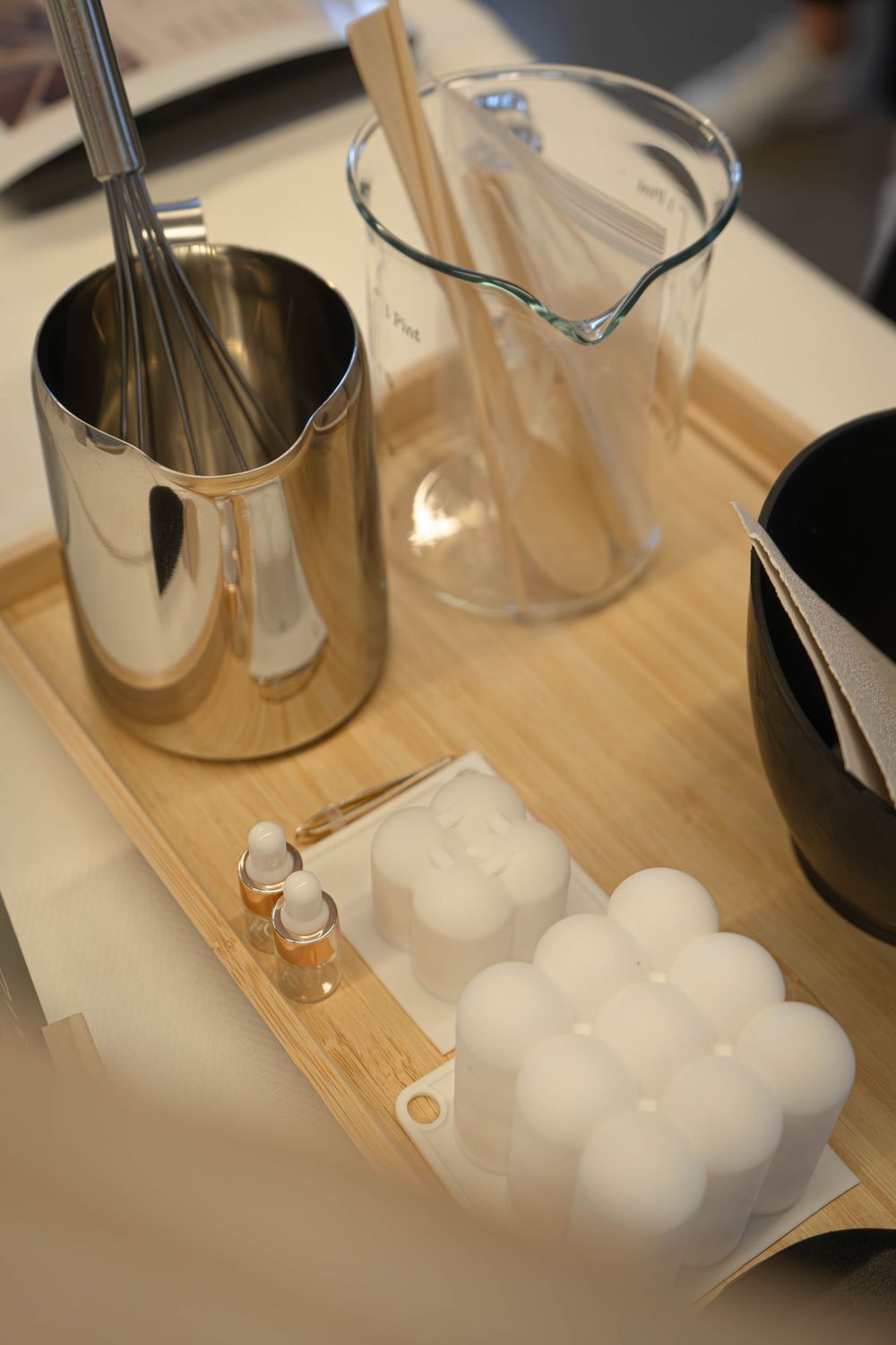 Candle making supplies on bamboo tray including metal pitcher, glass beaker, bubble mold, wax spheres, and fragrance oil.
