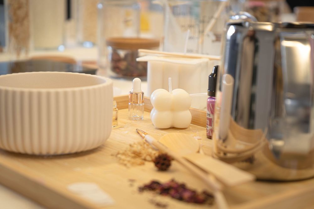 Candle making supplies on wooden table with white mold, bowl, dried flowers, and silver kettle.