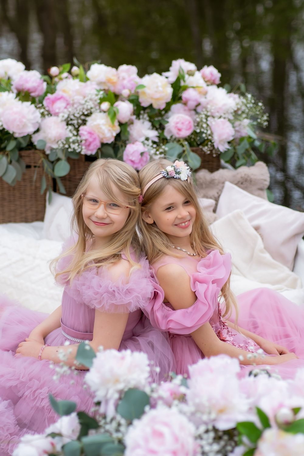 Two young children in pink tulle dresses pose among delicate pink roses and floral decorations in a dreamy garden setting.