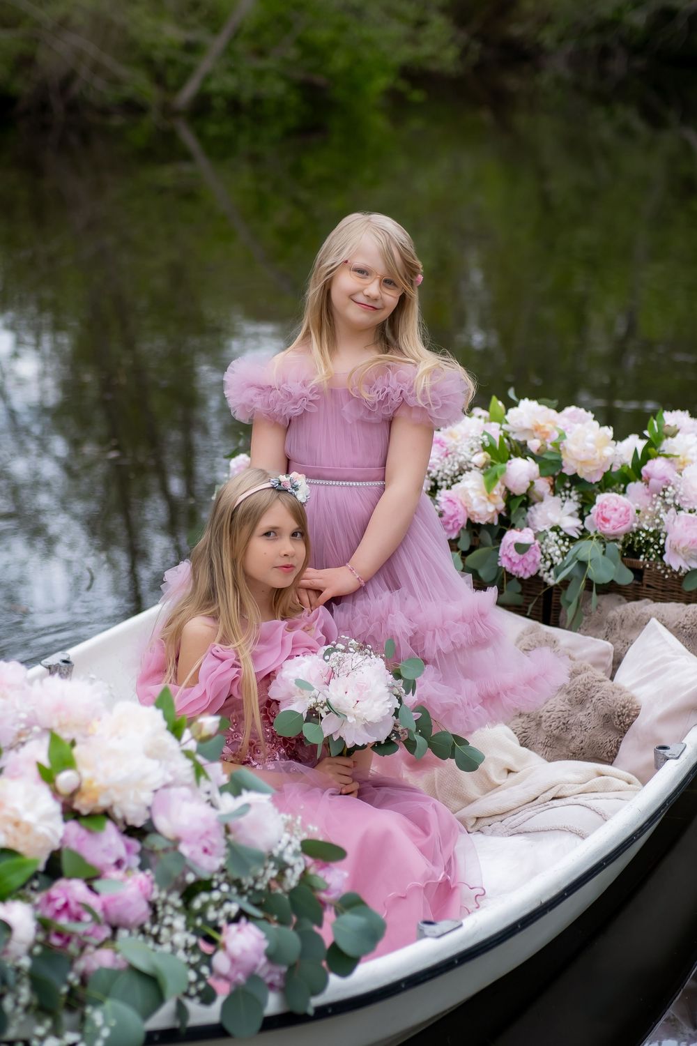 Two people in pink dresses sit in a white boat adorned with pink and white floral arrangements near a tranquil lake.