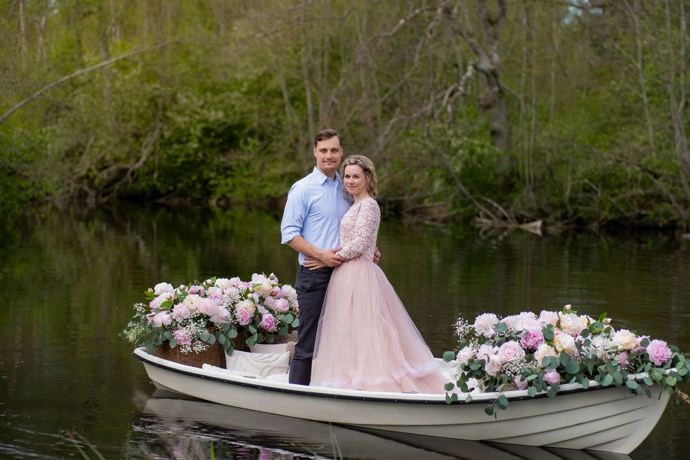 A romantic couple sits in a vintage rowboat decorated with pink and white floral arrangements on a serene lake.