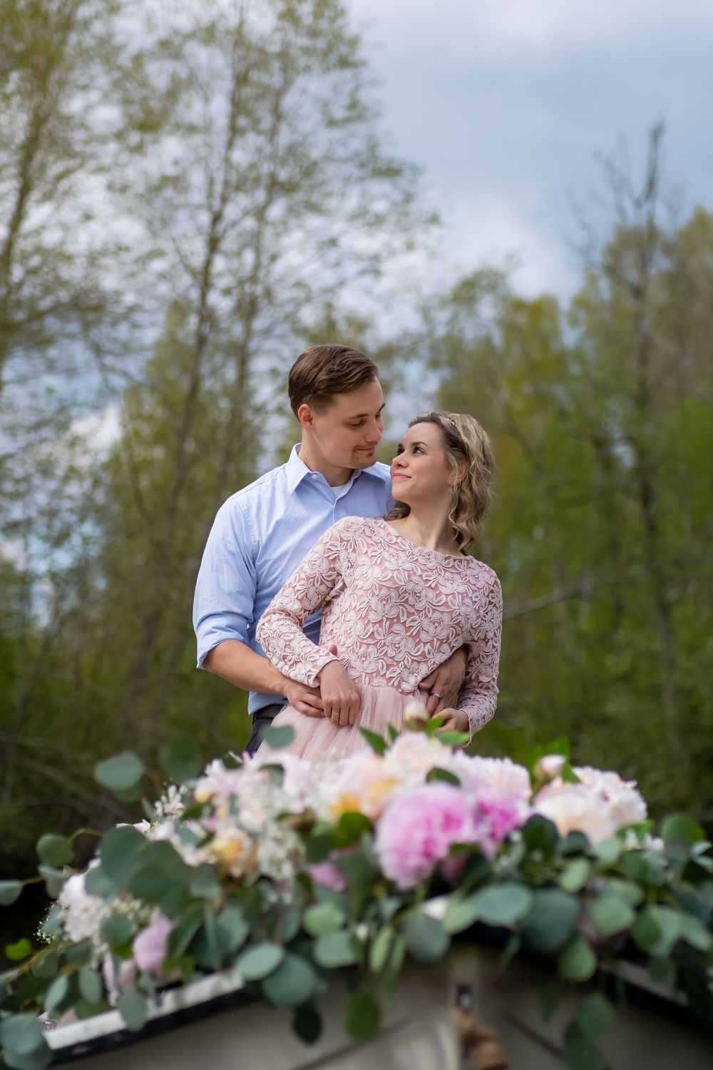 A romantic outdoor engagement photo shoot with floral decorations in the foreground and green trees in the background.
