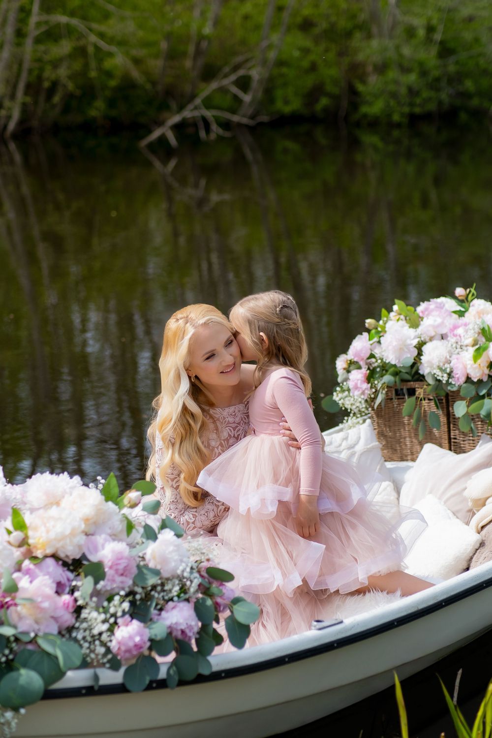A romantic couple in pink attire share tender moments while sitting in a flower-adorned boat on a tranquil lake.