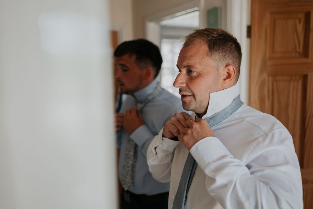 Groom adjusting his grey tie in front of a mirror while getting ready for his wedding day.
