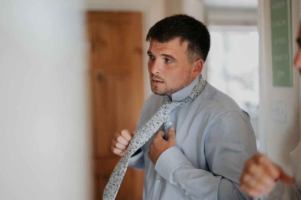 Groomsman in a light blue shirt tying his patterned tie while preparing for a wedding ceremony.