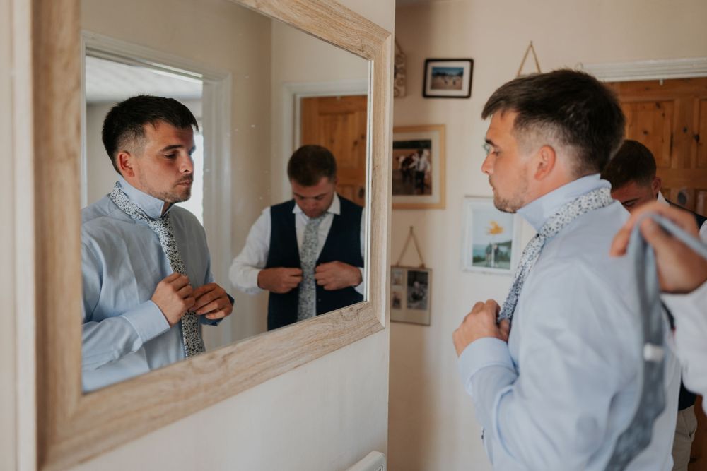 Two groomsmen in light blue shirts adjusting their ties while getting ready in front of a wooden mirror.