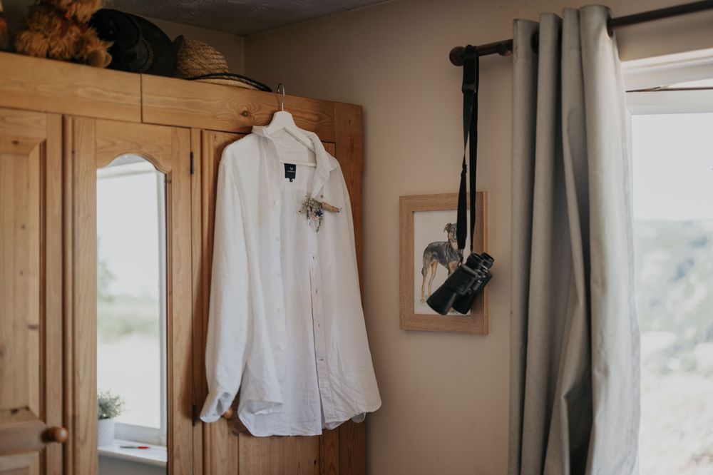 White linen groom shirt with boutonniere hanging on a wooden wardrobe near a window on wedding day.
