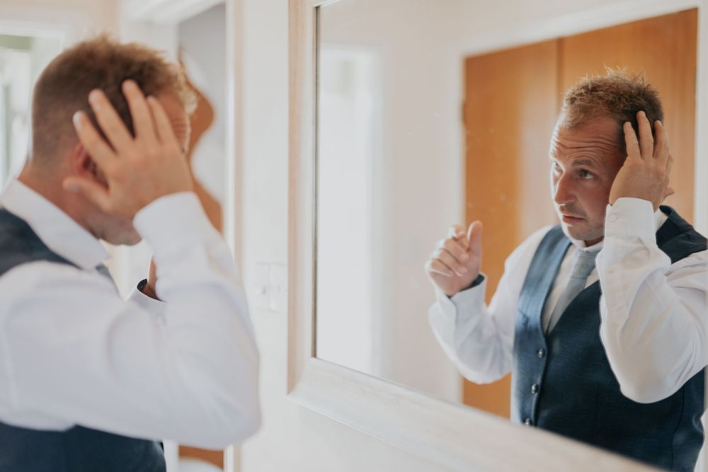 Groom in a navy vest and white shirt checking his hair in a mirror before his wedding ceremony.