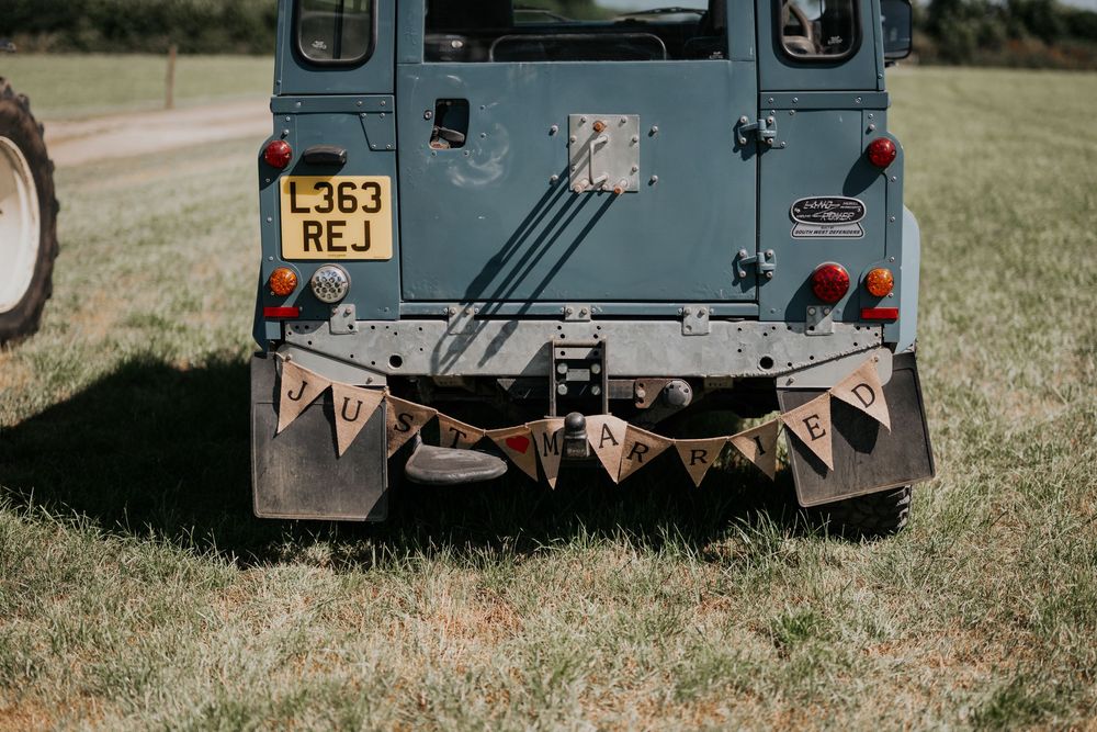 Rear view of a vintage Land Rover Defender with a rustic burlap Just Married bunting banner at a wedding.