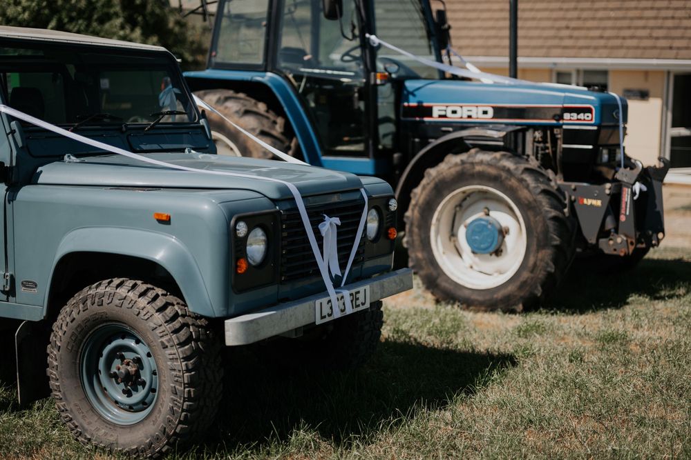 A classic Land Rover Defender with white wedding ribbon parked beside a blue Ford tractor on a sunny day.