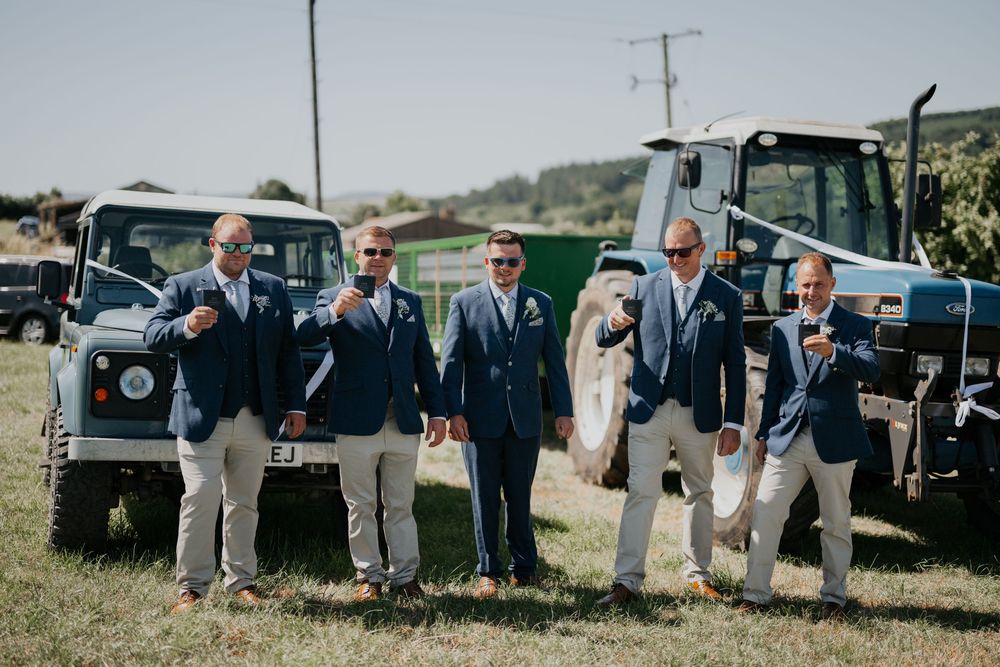 Groomsmen in navy suits holding drinks pose in front of Land Rover Defenders and a tractor at a farm wedding.