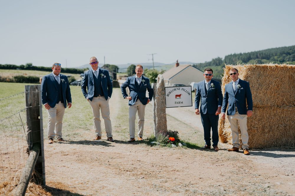 Five groomsmen in navy suits stand near a farm entrance sign with hay bales on a sunny wedding day.