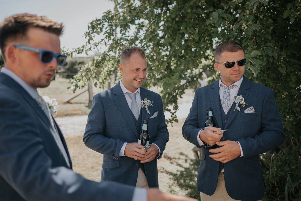 Three groomsmen in navy suits and sunglasses enjoying drinks outdoors under a tree at a summer wedding.