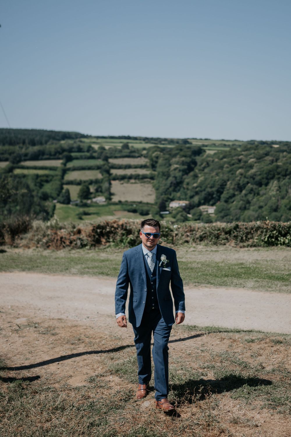 Dapper groom in navy suit and sunglasses walks along a gravel path with scenic green countryside behind him.