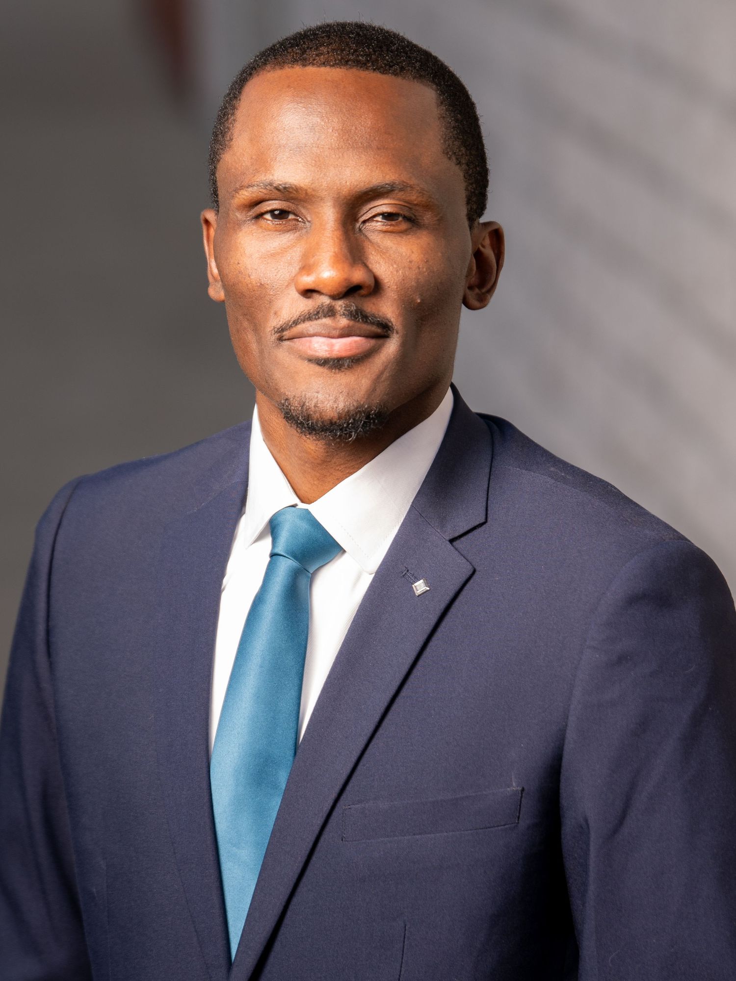 Professional portrait of someone in a navy suit and blue tie against a gray background.