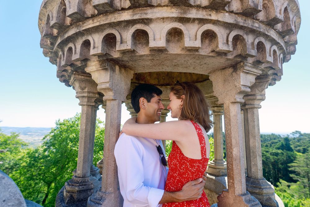 A couple embraces under an ornate stone gazebo surrounded by lush greenery and scenic views.