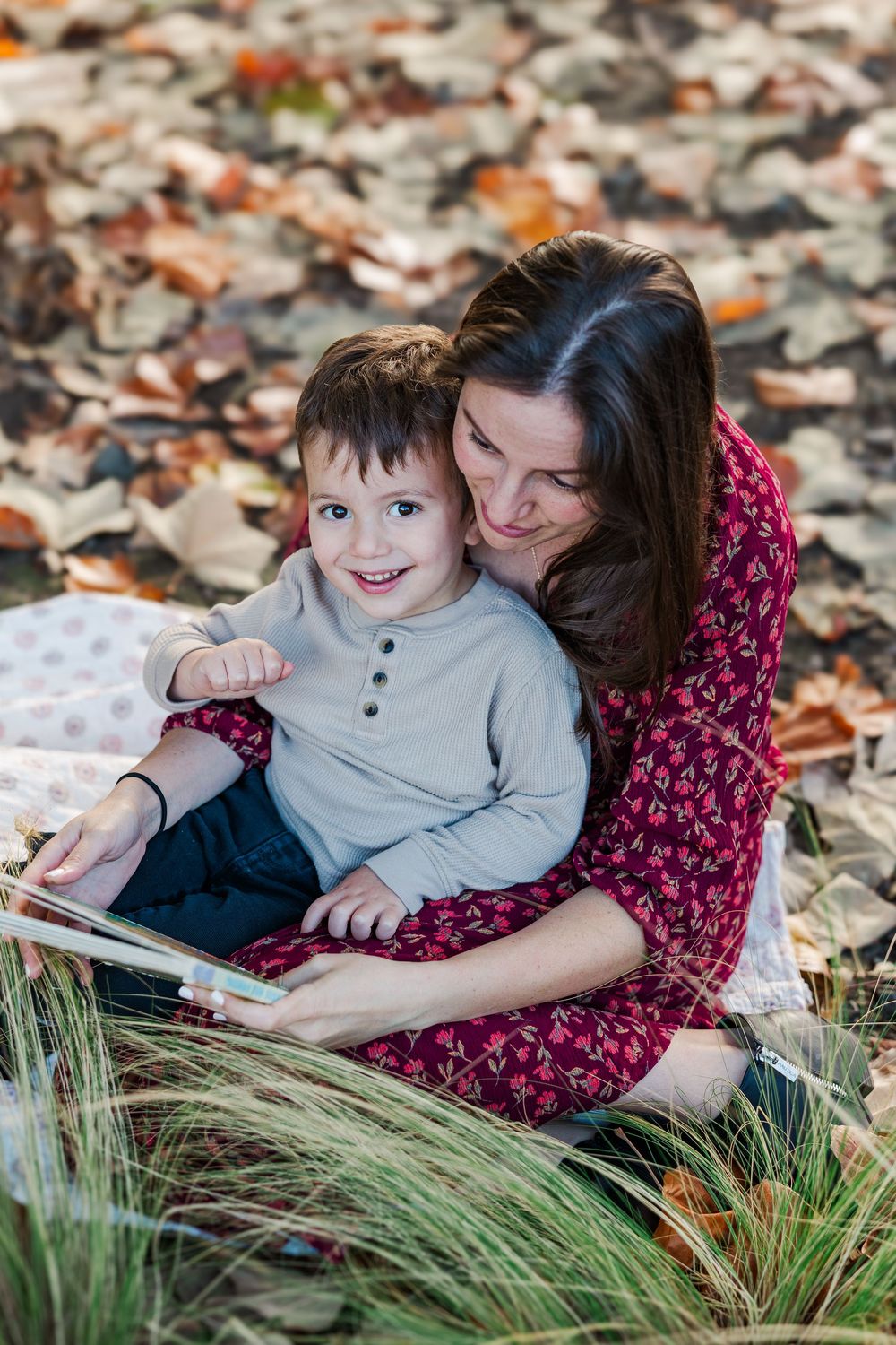 A cozy autumn scene with fallen leaves as two people snuggle together on the ground near tall grass.