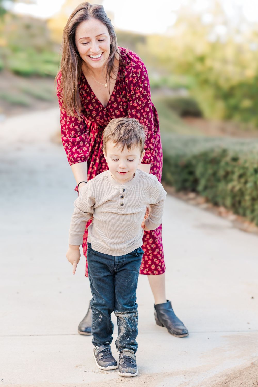 A toddler in beige sweater and jeans plays outdoors while accompanied by someone in a red polka dot top.
