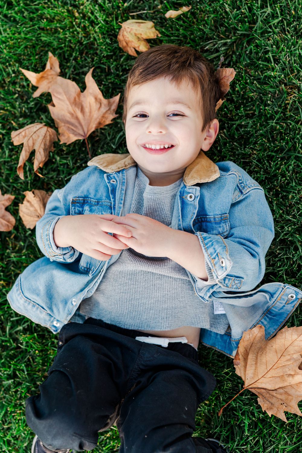 A child lying on green grass surrounded by autumn leaves wearing denim and gray clothing.
