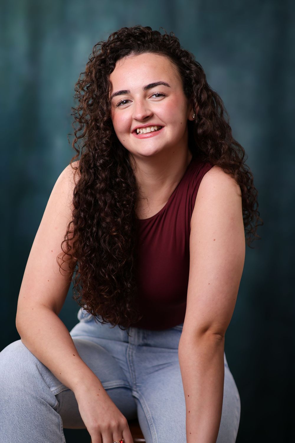 Portrait of someone with long curly dark hair wearing a burgundy sleeveless top and light jeans against a teal background.