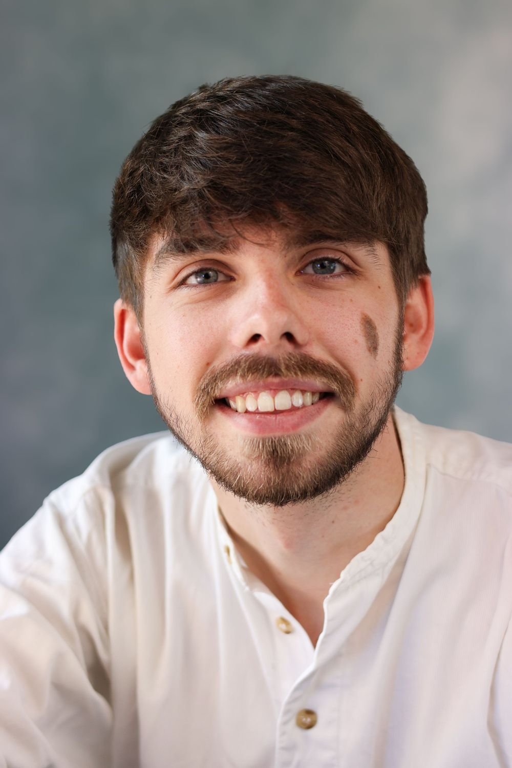 Portrait of person with dark hair and beard wearing white henley shirt against light background.