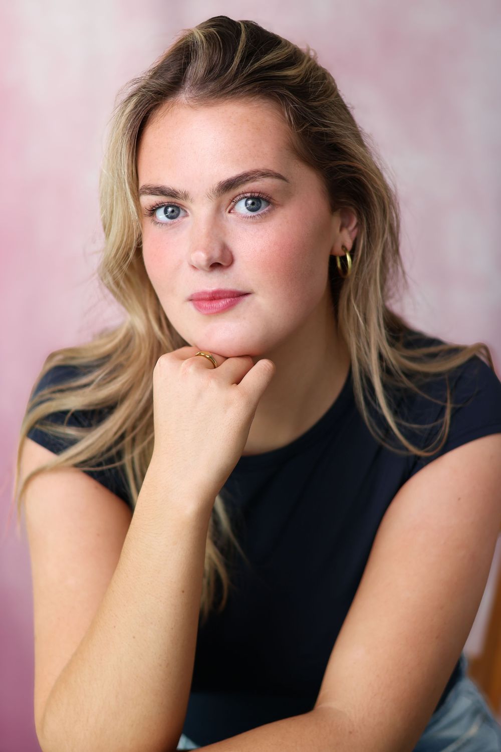 Portrait of a young woman in a black sleeveless top with blonde hair against a pink background.