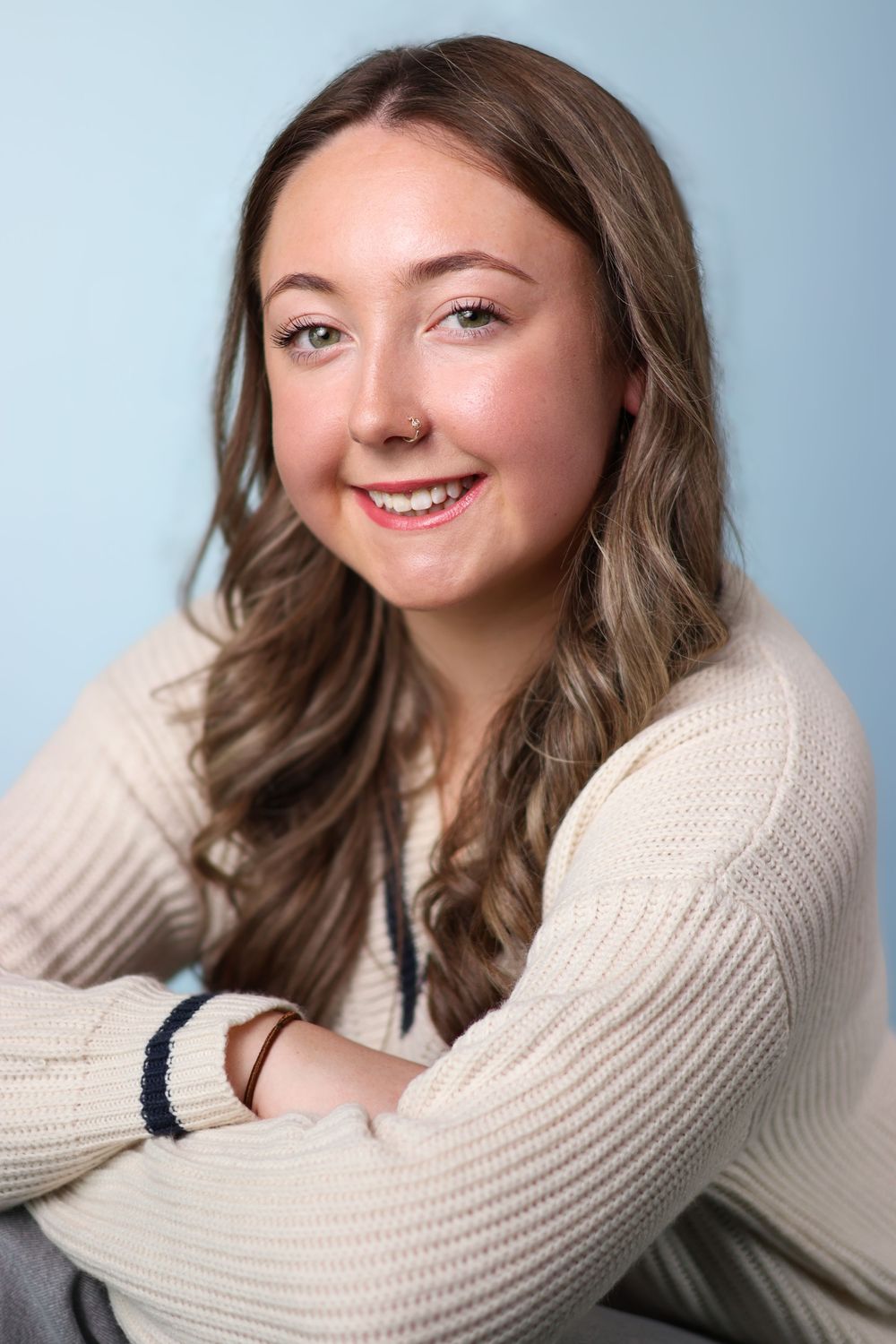 Professional portrait of someone with wavy brown hair wearing a cream sweater against a blue background, smiling warmly.