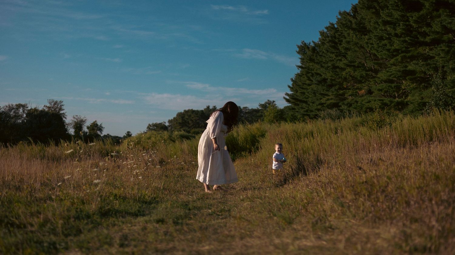 A couple walks together through a scenic meadow with tall grass and trees under a bright blue summer sky.