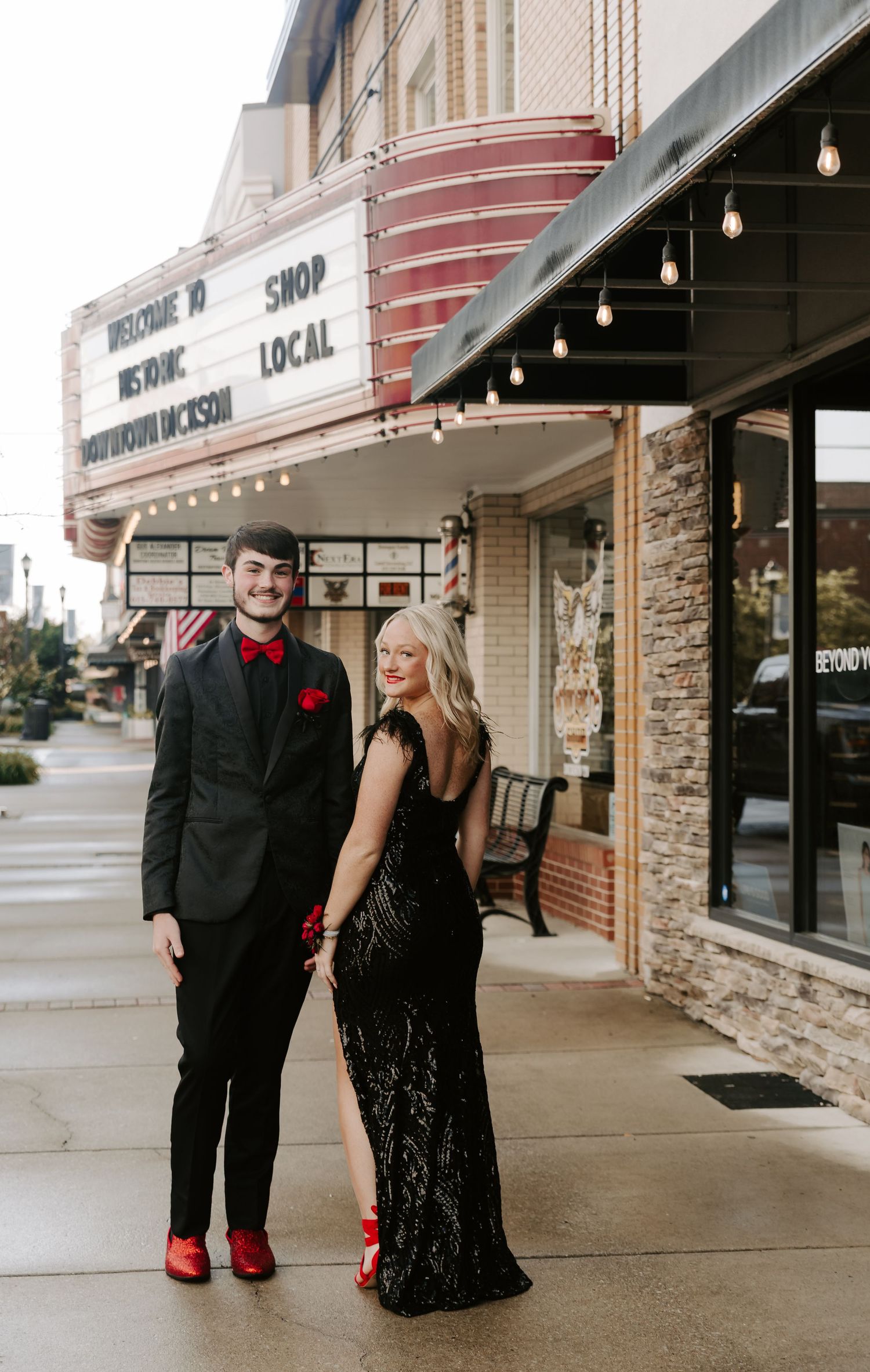 A couple dressed in formal attire poses together outside a local shop.