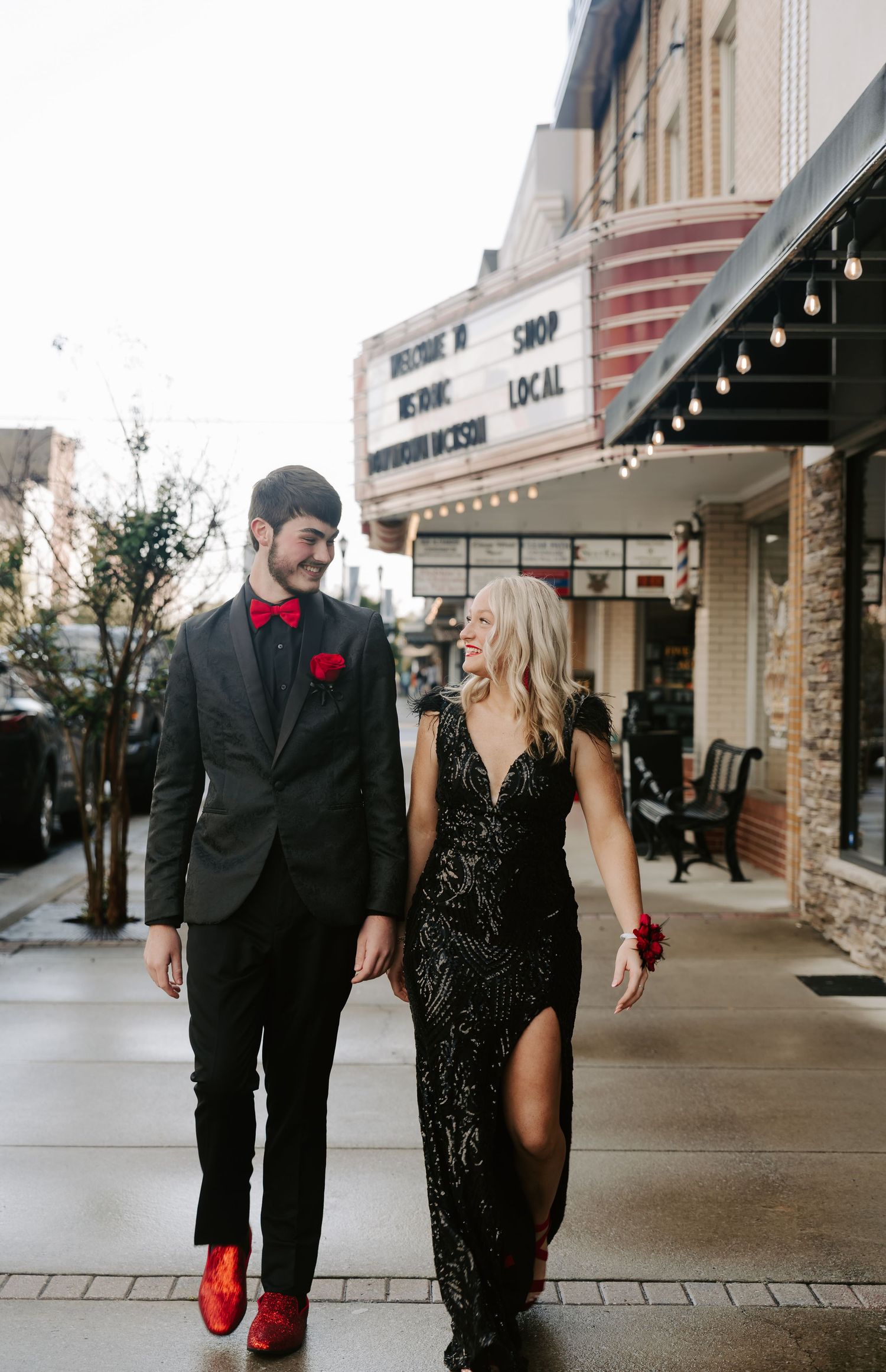 A couple in black formal wear strolls along a storefront sidewalk.