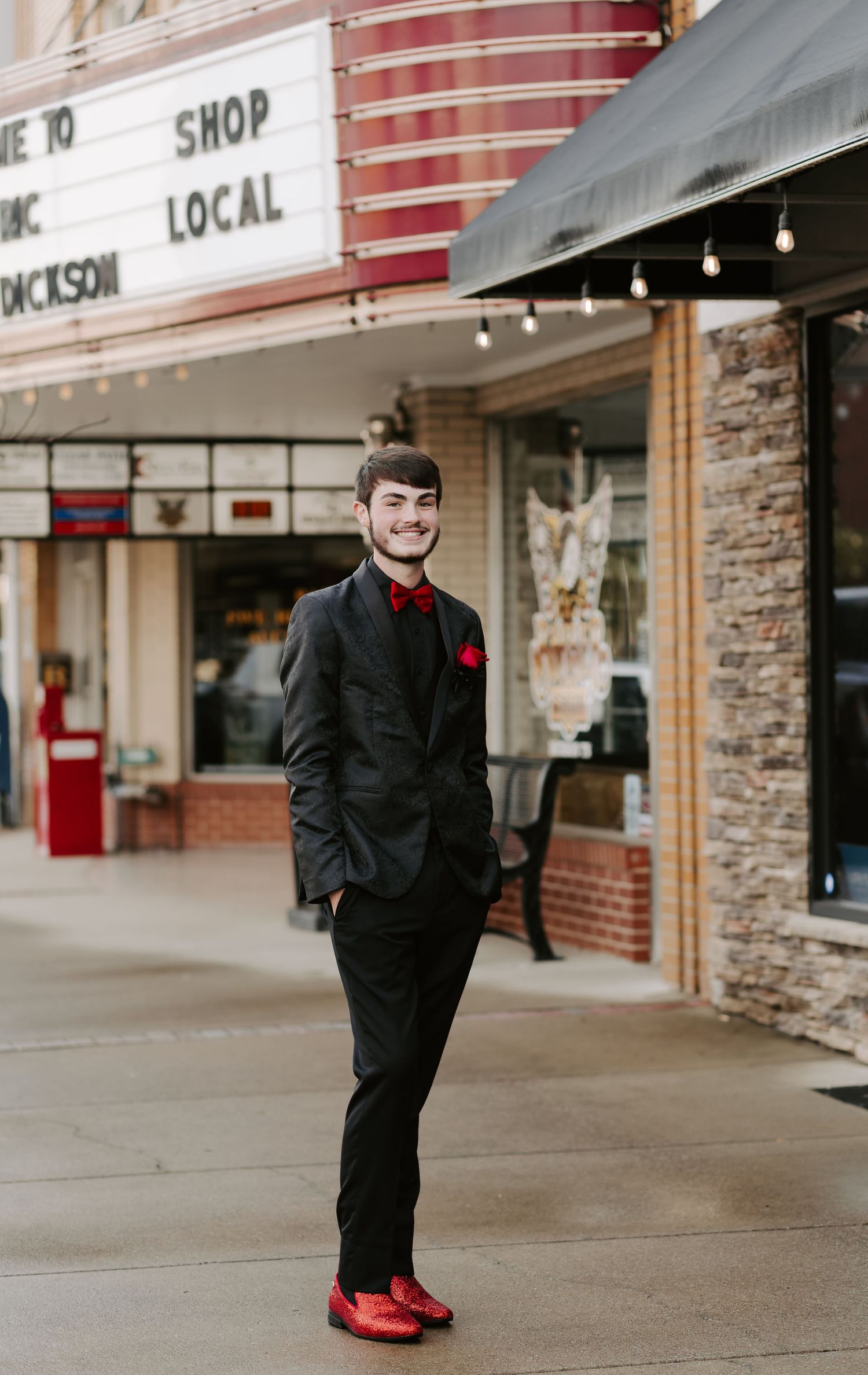 A person in a black suit and red shoes poses confidently outside a local shop.