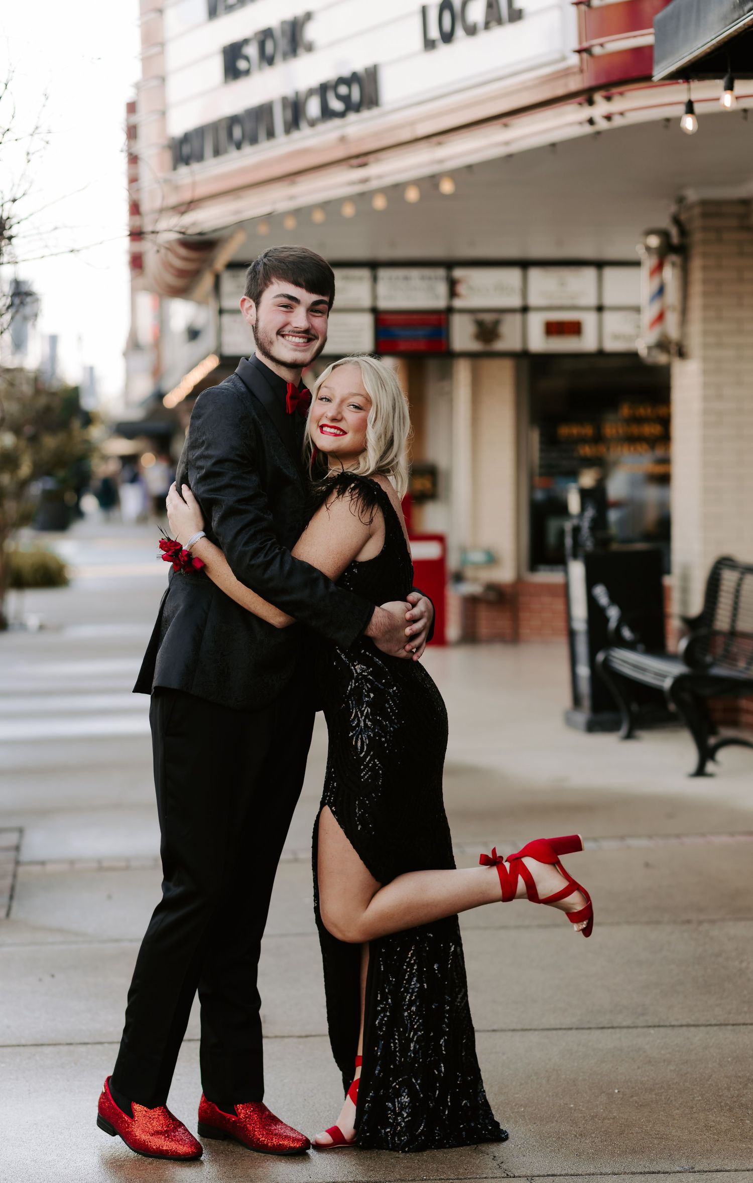 A couple in black attire with red accents poses playfully on a sidewalk.