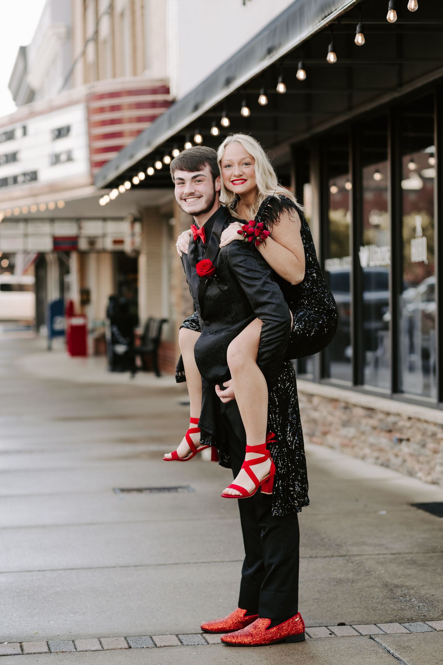 A couple in formal attire poses playfully on a city sidewalk with red accents in their outfits.