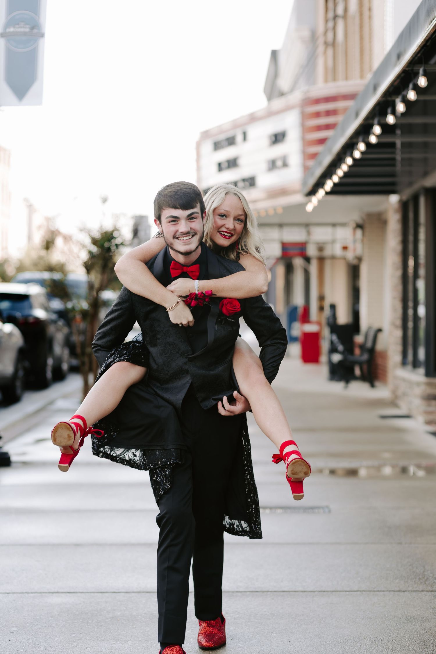 A couple shares a romantic moment in front of a storefront wearing black formal wear with red details.