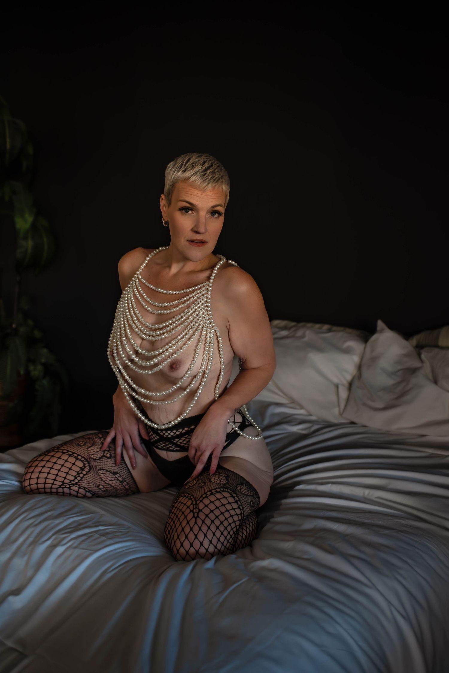 Boudoir photography of a woman wearing pearls, on her knees on a bed in a moody, intimate room in Salt Lake,