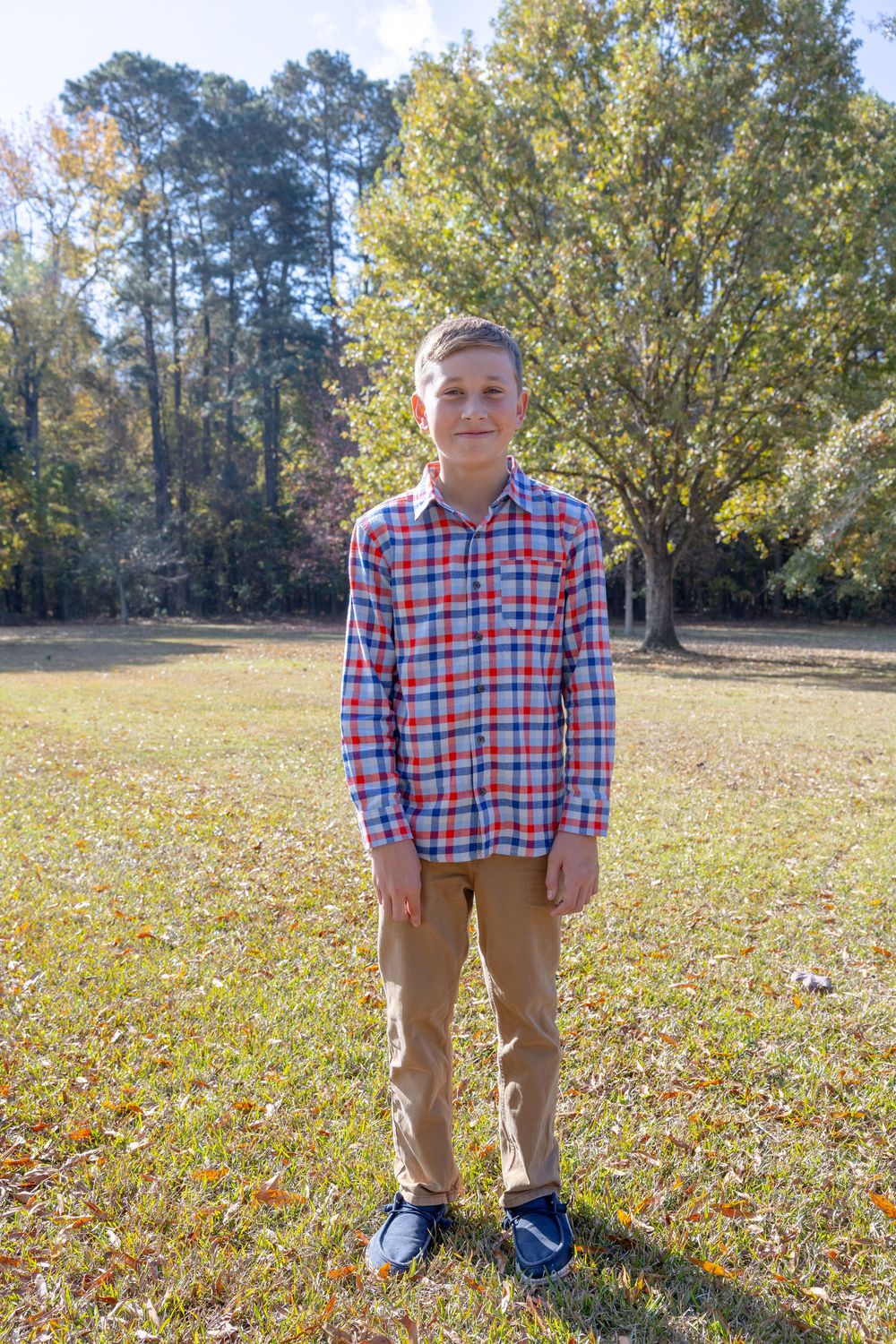 Person in plaid shirt and khakis stands on fallen leaves in a park setting during autumn.