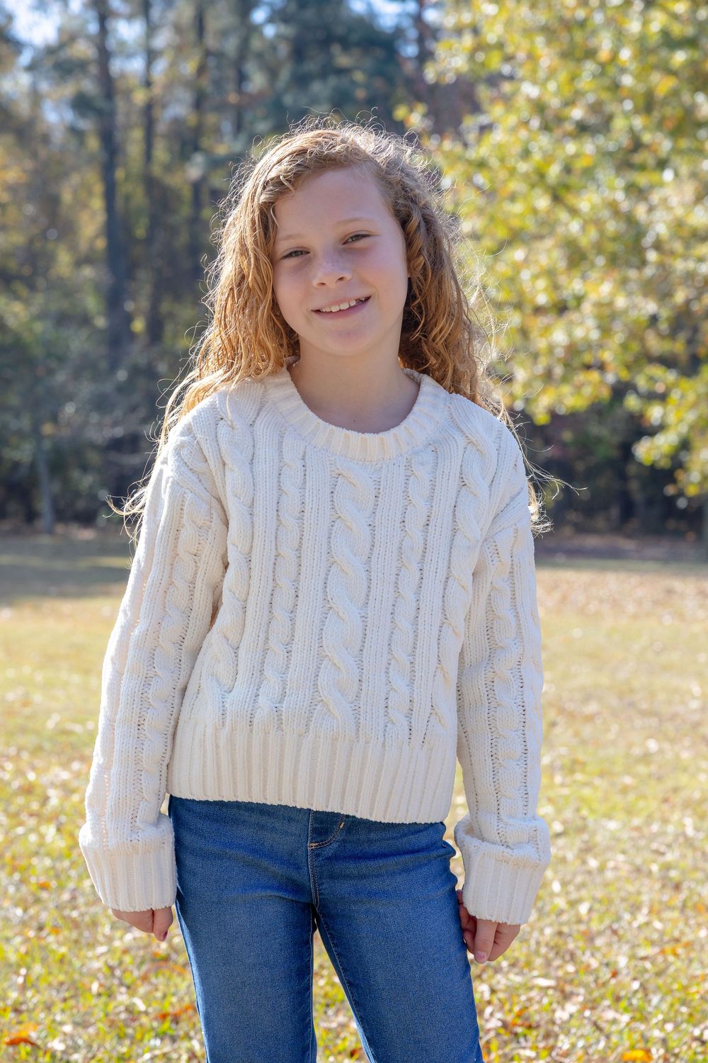 Young person wearing a white cable knit sweater and jeans poses outdoors on a sunny autumn day.