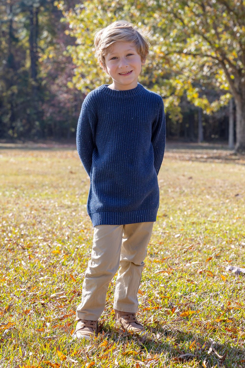 A child in a navy blue sweater and khaki pants stands in a sunlit field with fall foliage.
