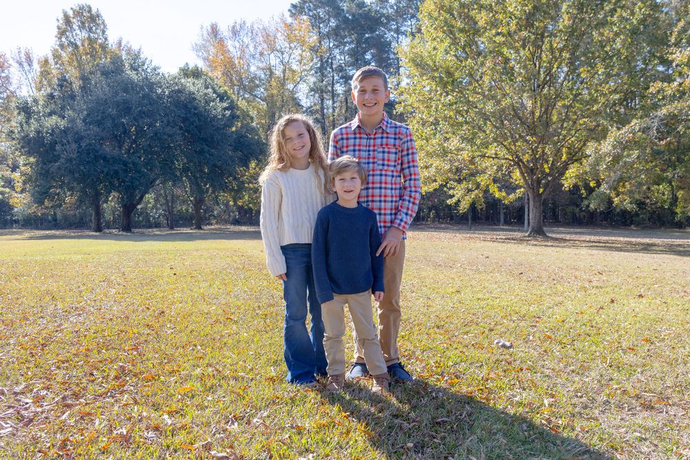 A family stands together in a scenic park setting with autumn trees in the background.