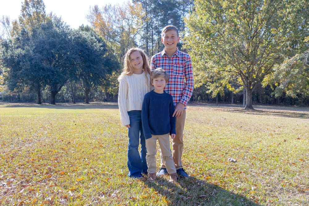 Three people stand together on grass with autumn trees in background during outdoor photo session.