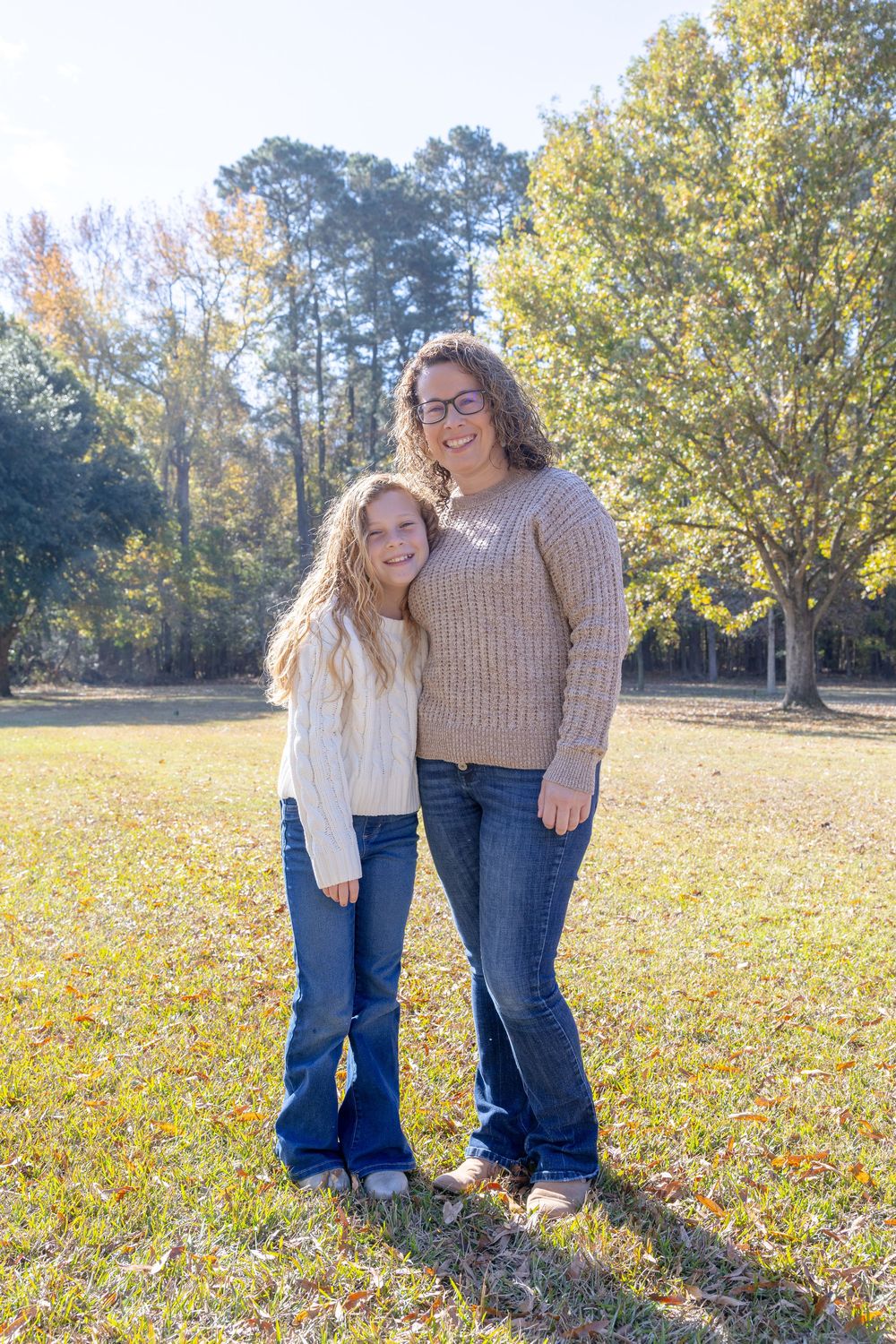 Two people in casual attire pose together on a grassy field with autumn trees in the background.