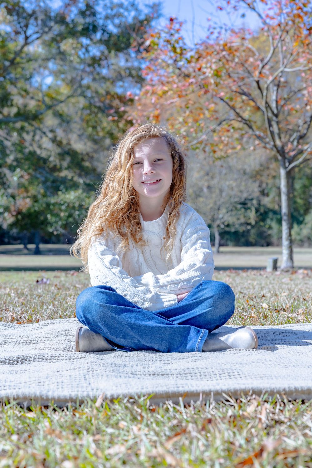 Person in white sweater sits cross-legged on blanket with fall foliage in background.