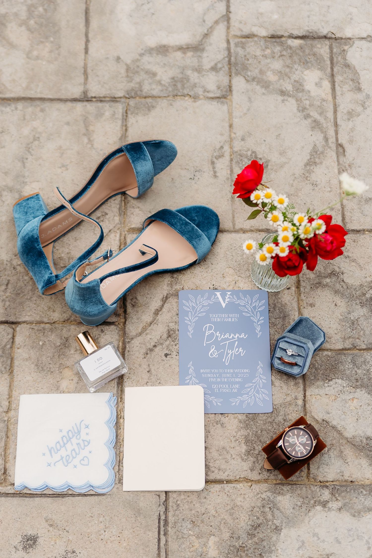 Wedding details flatlay with blue suede shoes, floral accents, and gray stationery on stone background.