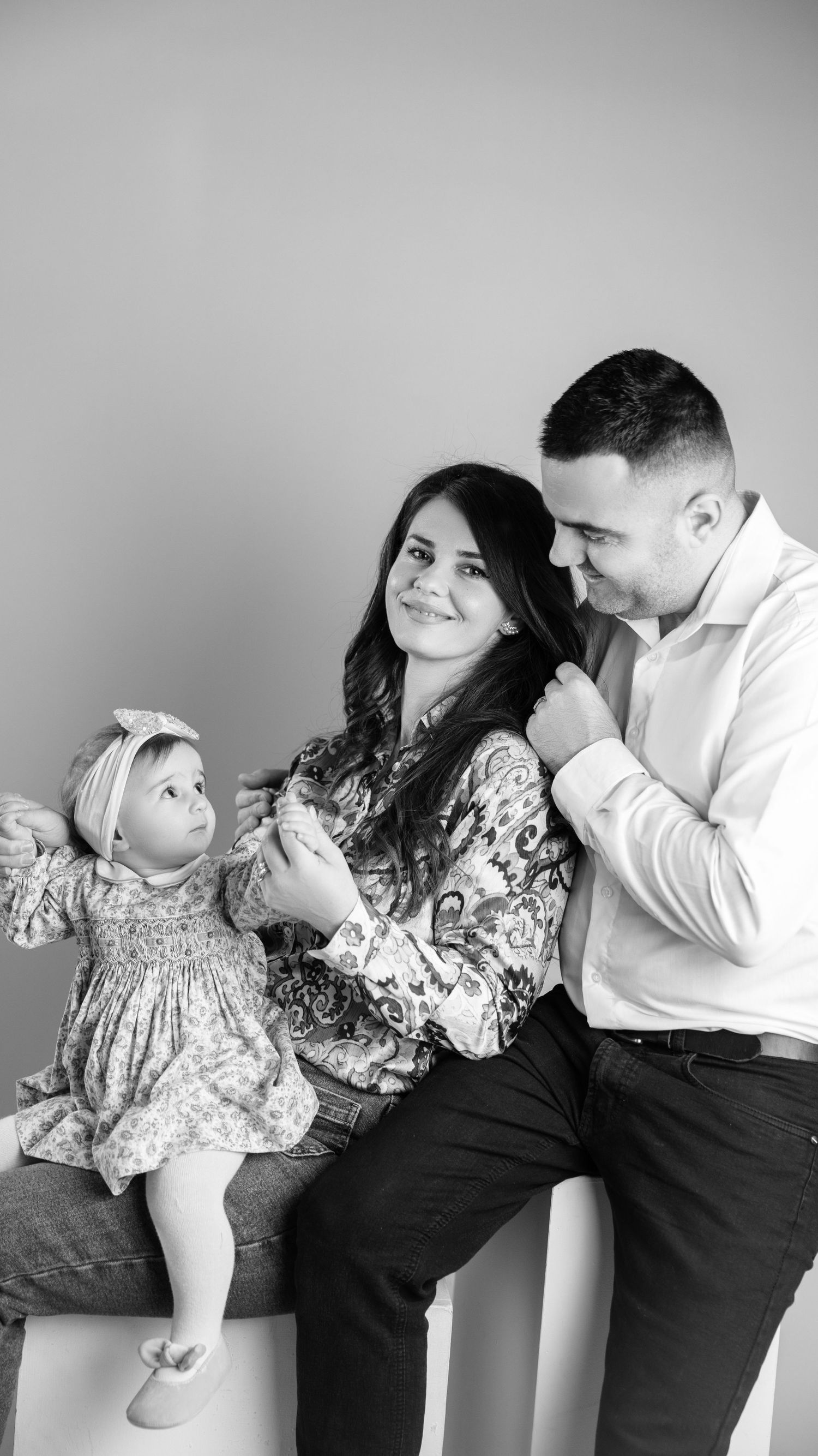 Black and white portrait of smiling couple with baby daughter in floral dress on studio bench.